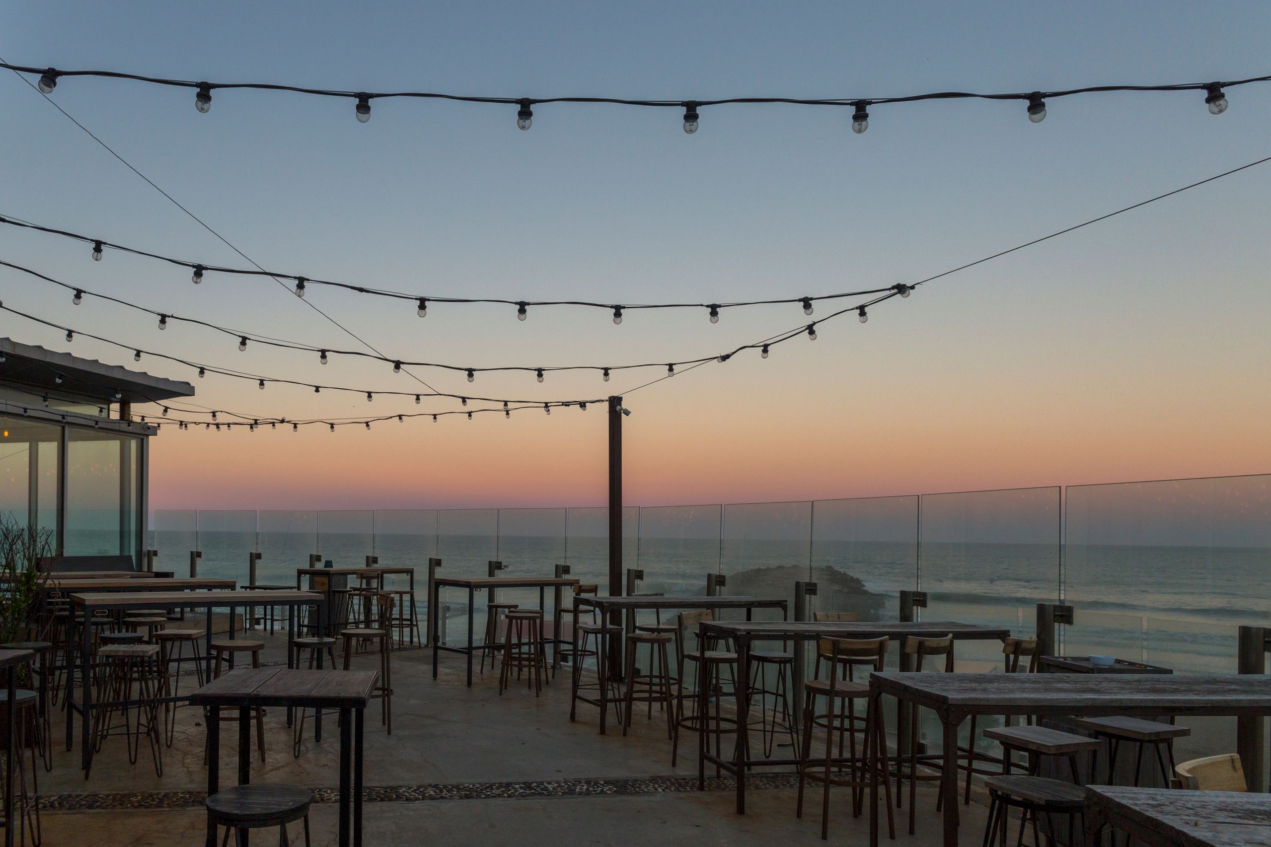 Exterior of Harbor Light Seafood Kitchen at dusk with string lights, blue umbrellas, outdoor seating, and harbor view